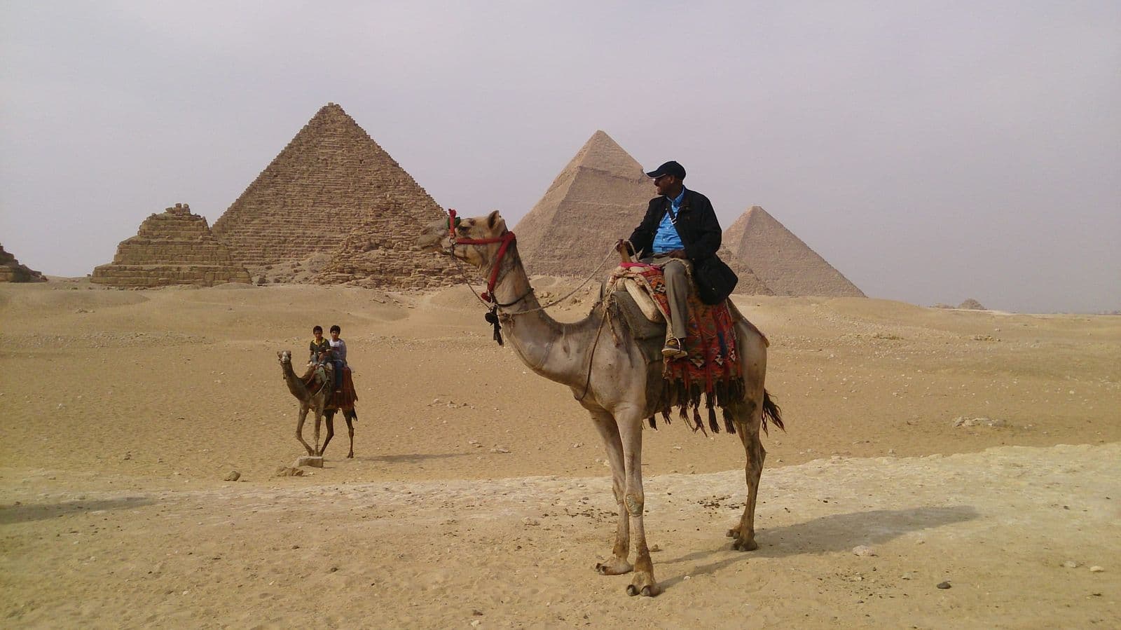 A camel rider on the sand with the three Pyramids of Giza behind