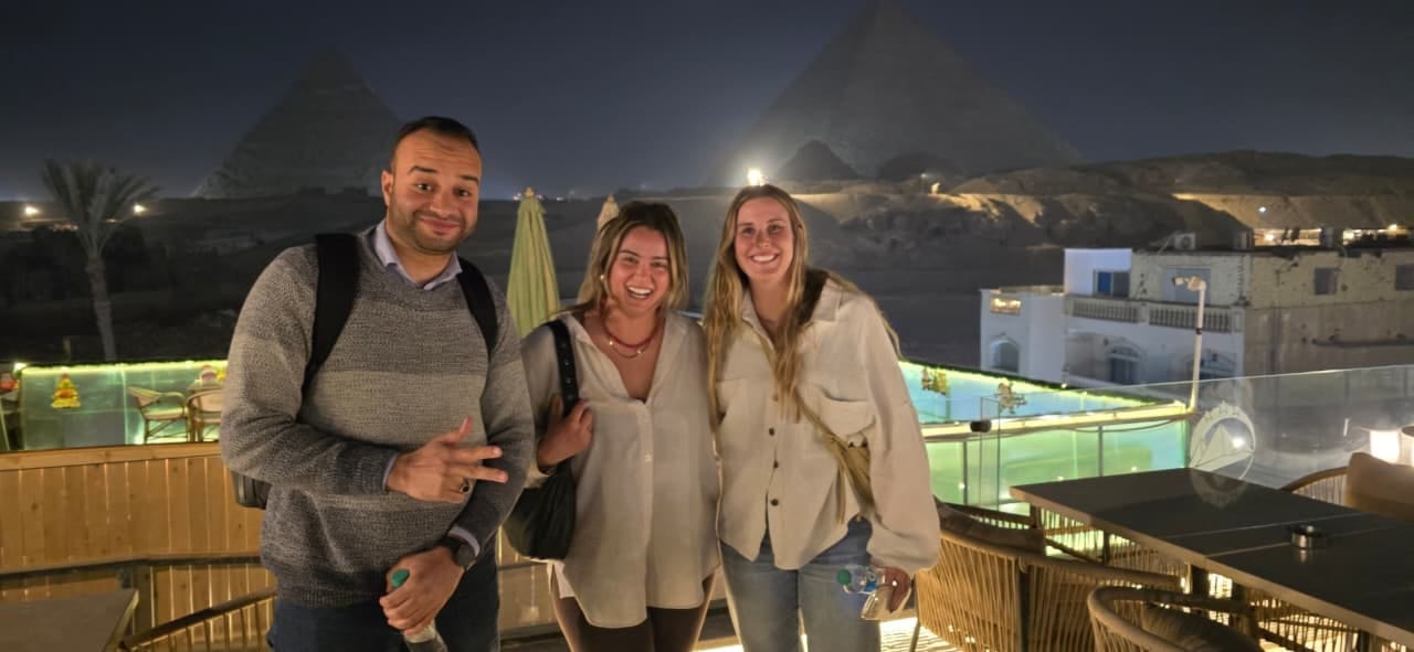 Tarek with two travelers on a rooftop at night, the lit pyramids of Giza behind