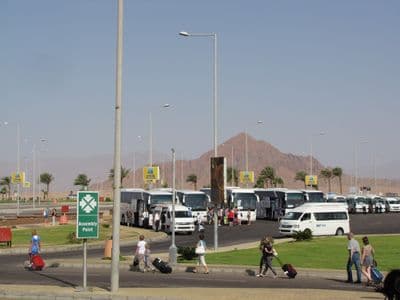Travelers with suitcases walking to waiting minibuses outside Sharm el-Sheikh airport, mountains behind